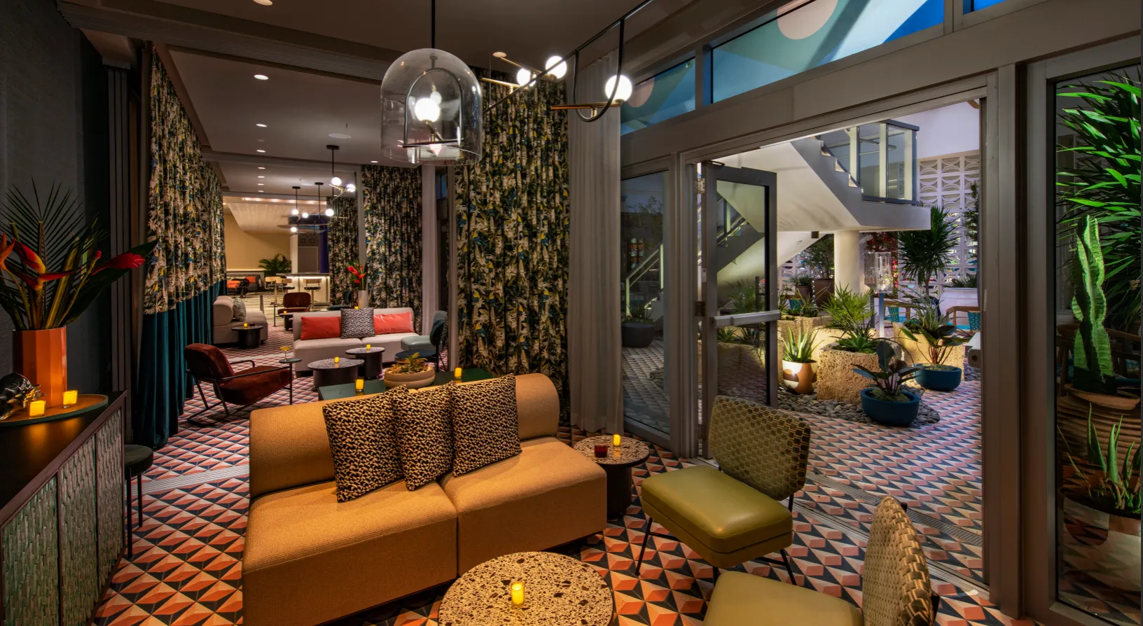 A stylish indoor lounge with patterned tile floors, modern furniture, warm lighting, and glass doors opening to a patio with plants and an outdoor staircase. The space feels cozy and inviting.