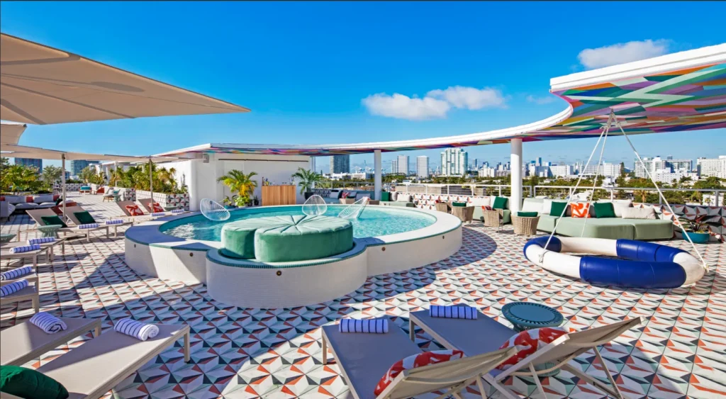 A colorful rooftop lounge features a round pool with green cushioned seats, striped lounge chairs, patterned tile flooring, umbrellas, and a city skyline in the background under a clear blue sky.