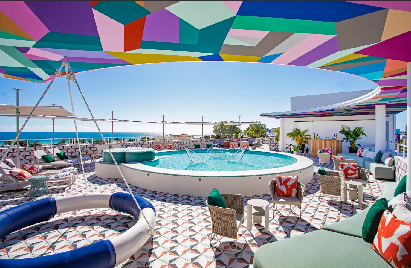 A vibrant rooftop lounge with a round pool, colorful geometric ceiling, patterned floor tiles, cozy seating, and ocean views under a clear blue sky.