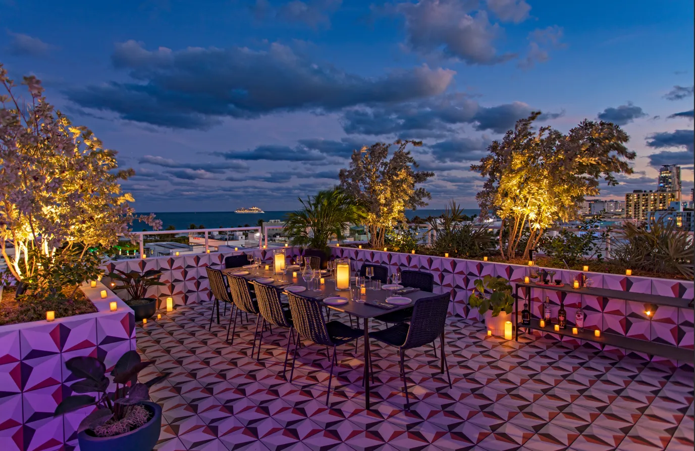 Rooftop dining area at dusk with geometric tile floor, modern black chairs around a table, glowing candles, lush plants, and a view of the ocean and city skyline under a partly cloudy sky.